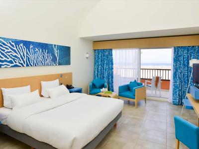 Bright hotel room with double bed, blue curtains, and balcony with sea view.