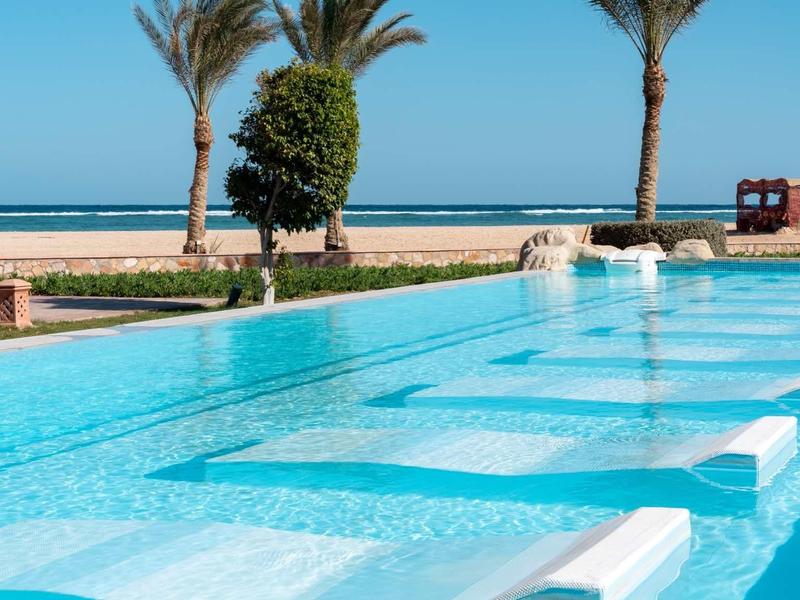 Clear pool with built-in loungers, palm trees, and blue sky by the sea.