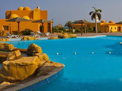 Großer blauer Hotelpool mit Felsen und gelben Gebäuden im Hintergrund bei klarem Himmel.