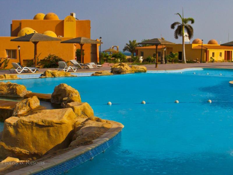 Piscine bleue avec rochers et bâtiments jaunes sous un ciel bleu clair.