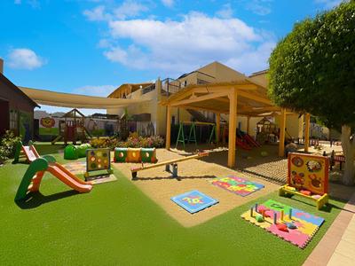 Kinderspielplatz mit Rutsche, Schaukeln, Spielhäusern und bunten Spielmatten unter blauem Himmel.