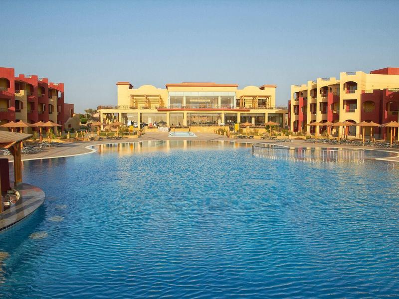 Großer blauer Pool mit roten Gebäuden und Sonnenschirmen unter klarem Himmel.