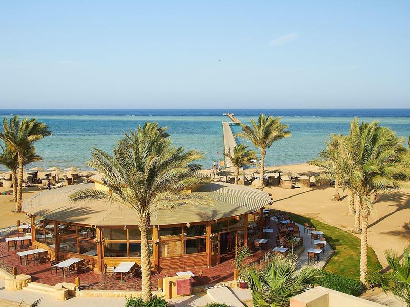 Strand mit Palmen, Rundbau mit Sitzplätzen und blauem Meer im Hintergrund bei klarem Himmel.