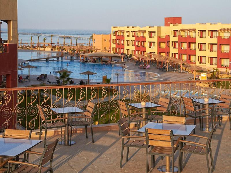 Balkon mit Stühlen und Tischen vor bunten Häusern und Wasser im warmen Abendlicht.