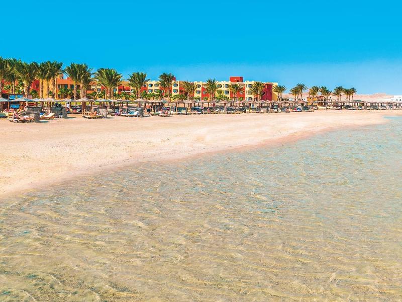 Breiter Sandstrand mit klarem Wasser, bunten Gebäuden und Palmen unter blauem Himmel.