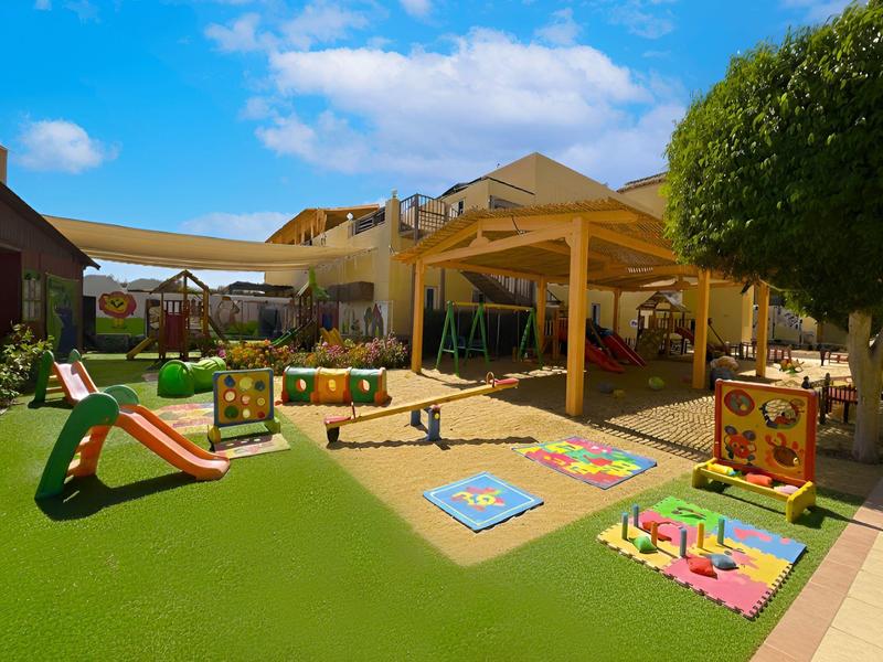 Kinderspielplatz mit Rutsche, Schaukeln, Spielhäusern und bunten Spielmatten unter blauem Himmel.