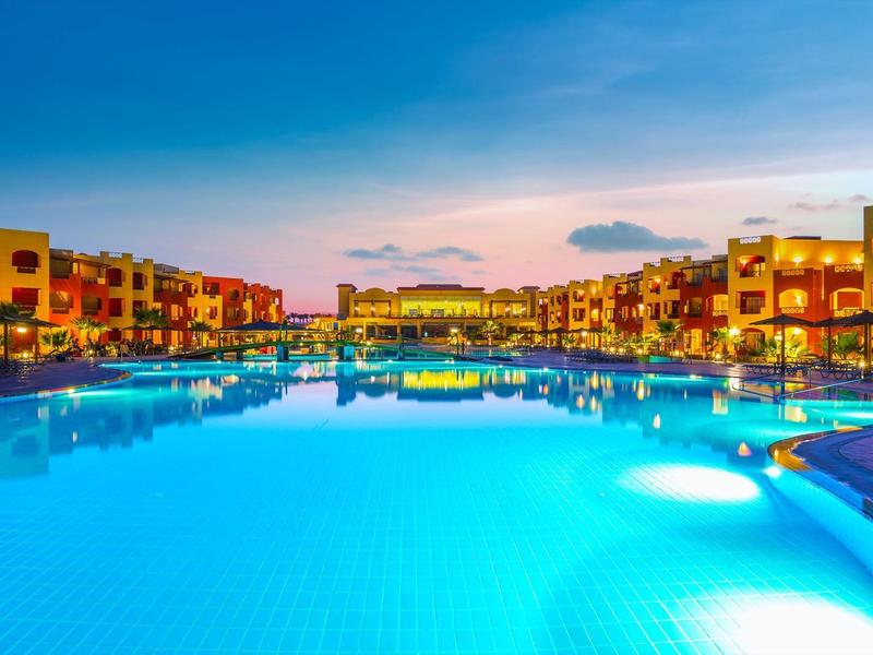 Großer beleuchteter Pool bei Sonnenuntergang, umgeben von Hotelgebäuden mit Balkonen.