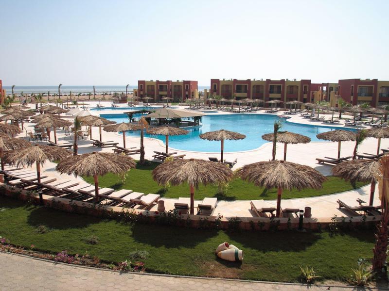 Großer, runder Pool mit vielen Liegen und Strohdächern, umgeben von roten Gebäuden und Palmen.