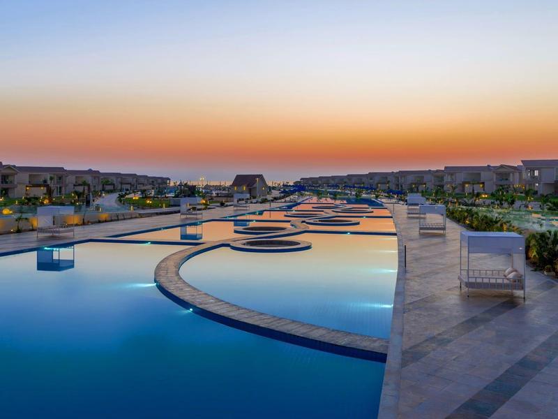 Piscine à débordement dans un hôtel au coucher du soleil avec chaises longues et paysage urbain.