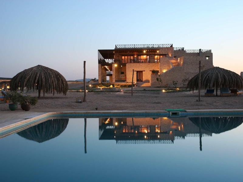 Luxuriöses Haus mit warmen Lichtern, Pool und zwei Strohdächern bei Dämmerung.