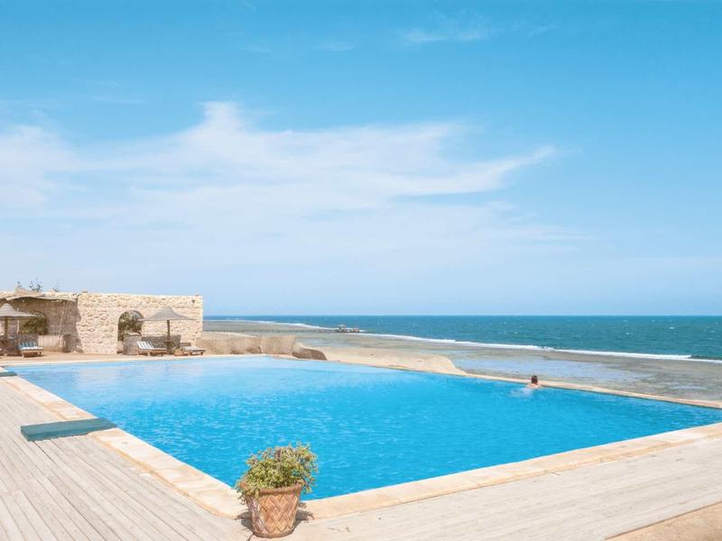 Infinity-Pool mit blauem Wasser, Holzdeck und Blick auf ruhiges Meer unter hellem Himmel.