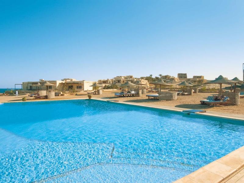 Großer Pool mit klarem Wasser und mehreren Liegen, im Hintergrund Häuser unter blauem Himmel.