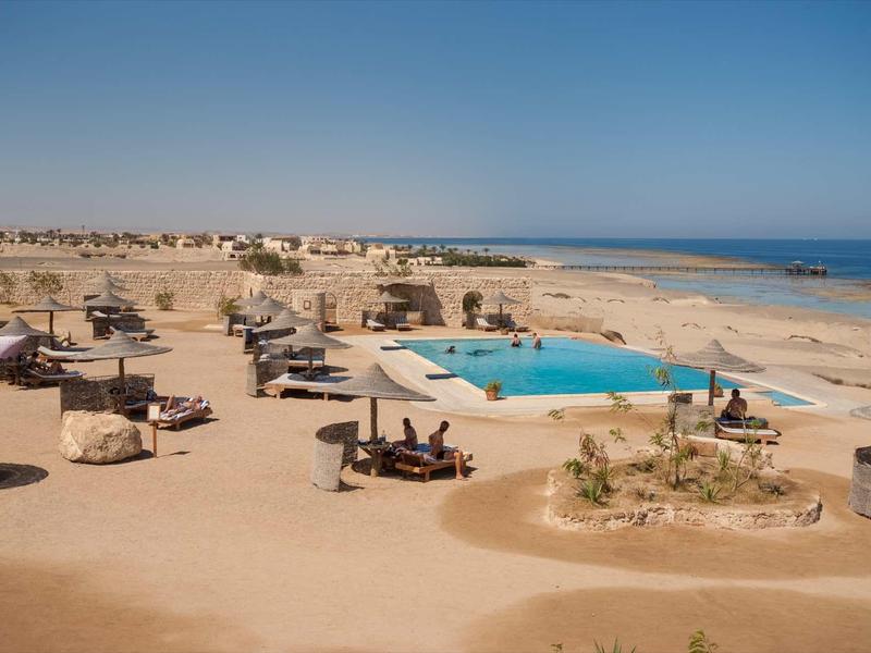 Strandresort mit Pool, Sonnenliegen und Sandhügeln an einem klaren, blauen Himmel.