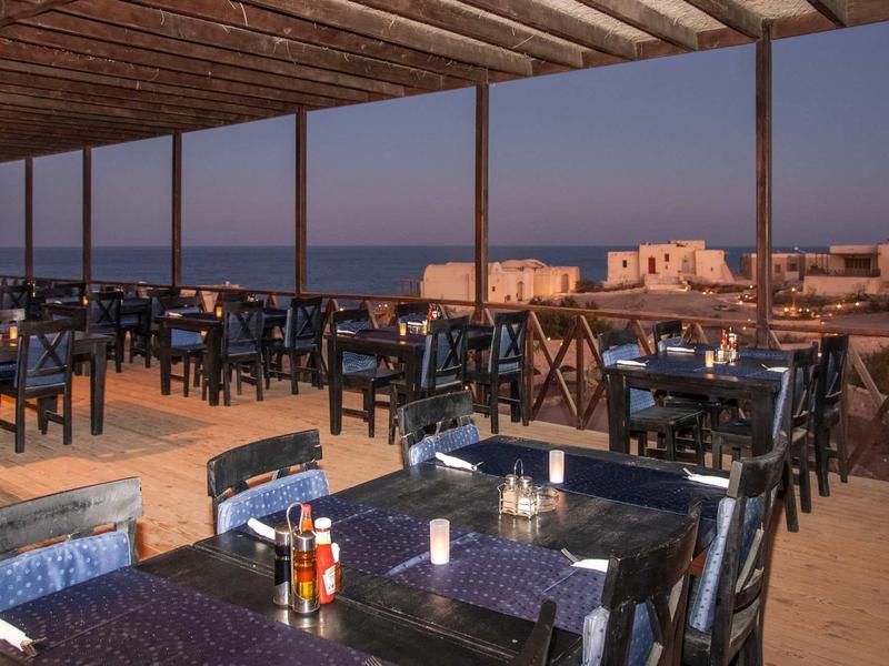 Restaurantterrasse mit blau lackierten Stühlen, Tischen und Blick auf weiße Häuser und Meer im Abendlicht.