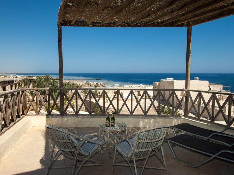 Balkon mit Stühlen und Tisch unter Holzdach, Blick auf Meer und Gebäude bei klarem Himmel.