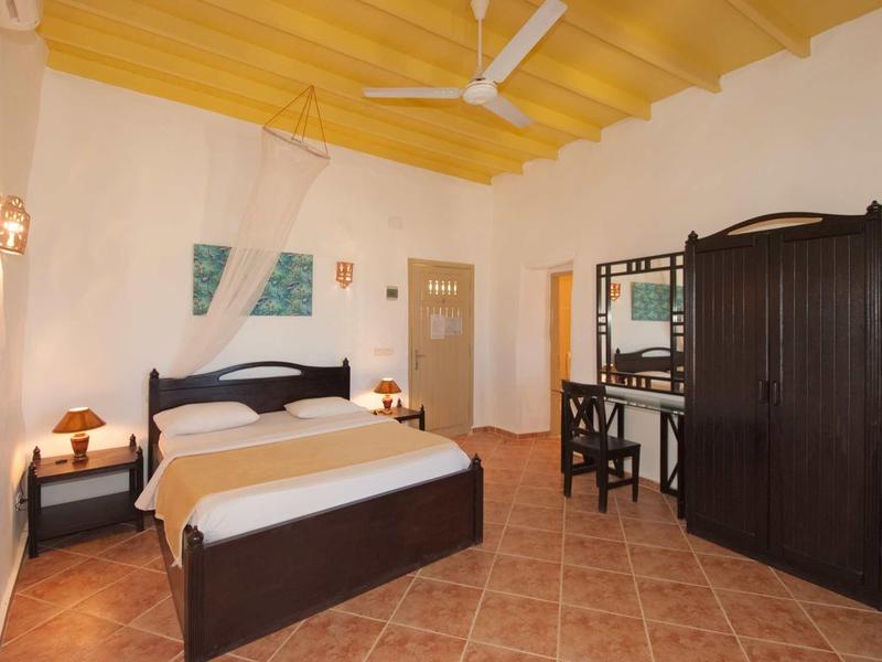 Helles Schlafzimmer mit gelbem Holzdeckenventilator, dunklem Holzmöbel und terrakottafarbenem Boden.