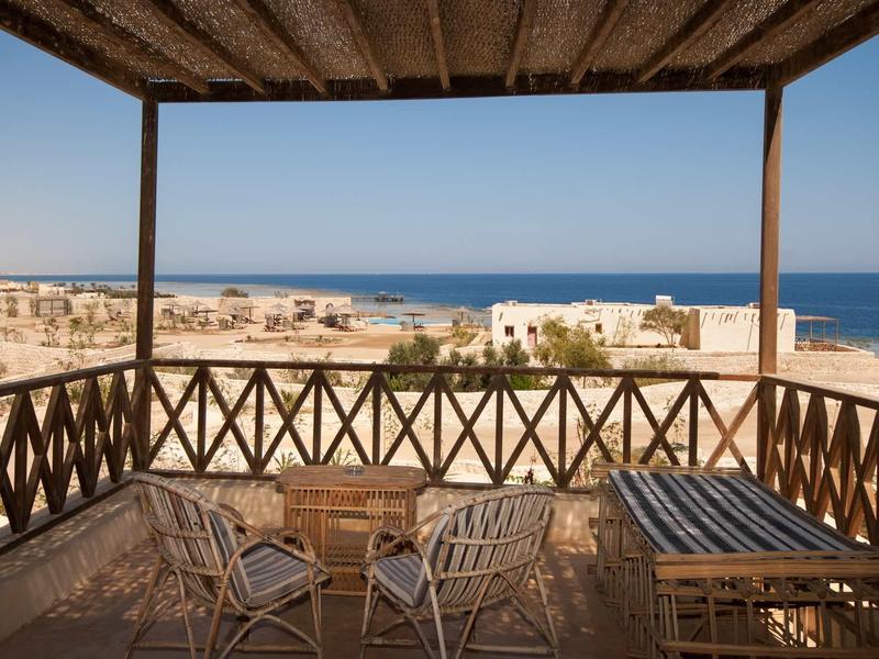 Überdachte Holzterrasse mit Tisch und Stühlen, Blick auf sandige Landschaft und blaues Meer.