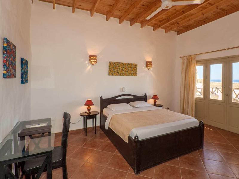 Schlafzimmer mit Holzbalkendecke, Doppelbett, Schreibtisch, zwei Nachttischen und großen Fenstern.