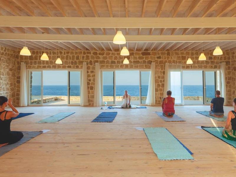 Innenraum mit großen Fenstern, durch die Meer und Himmel sichtbar sind, Menschen machen Yoga auf Matten.