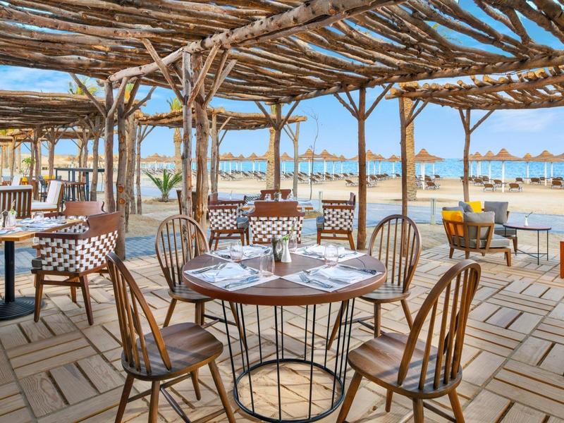Terrasse de restaurant en bois avec vue sur la plage et parasols sous un ciel bleu clair.