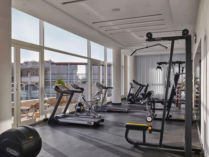 Salle de sport lumineuse avec tapis roulants et machines, grandes fenêtres avec vue extérieure.