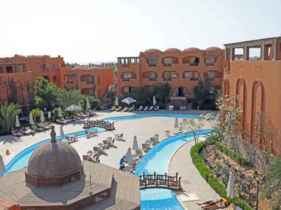 Hotelanlage mit rotem Sandstein, Pool und Sonnenliegen bei sonnigem Himmel.