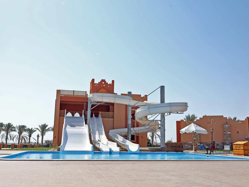 Wasserpark mit großen Wasserrutschen, flachem Pool und orangefarbenen Gebäuden im Hintergrund.