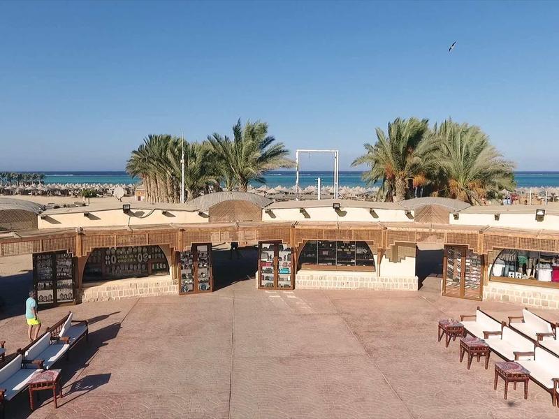 Offene Marktplatzfläche mit Schattendach, Sitzbänken und palmenbewachsenem Hintergrund am Meer.