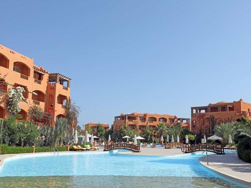 Großer Pool mit bläulichem Wasser vor orangefarbenen Apartmentgebäuden unter klarem Himmel.