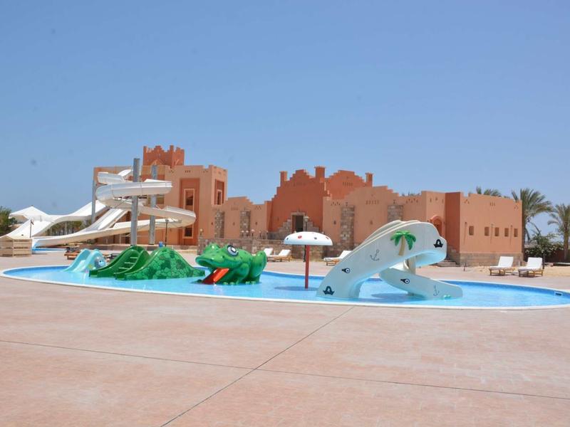 Freizeitbad mit Wasserrutschen, Planschbecken, Sonnenschirmen und rotbraunen Gebäuden im Hintergrund.