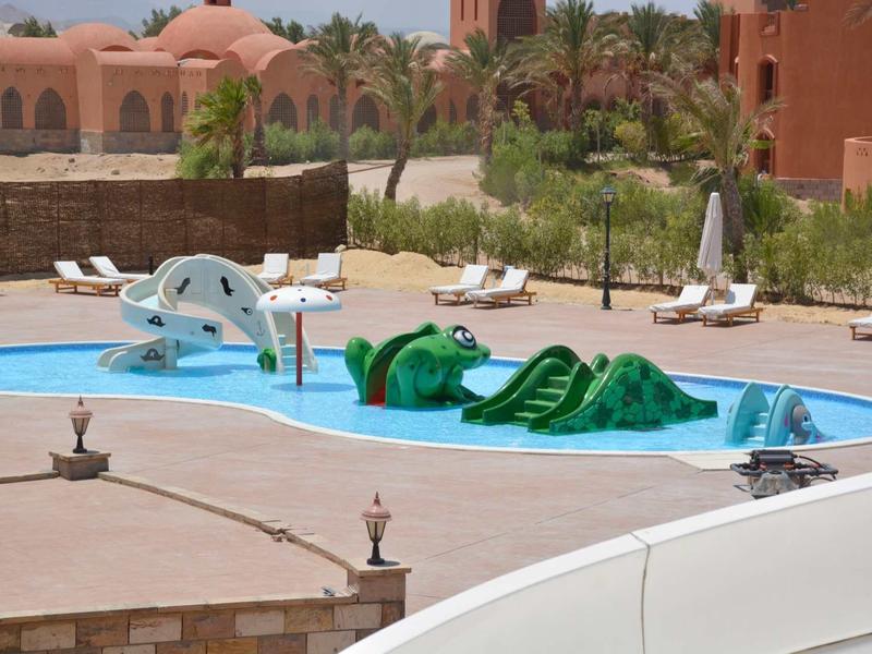 Kinderplanschbecken mit Wasserrutsche und Krokodilfigur, umgeben von Liegestühlen und Palmen.