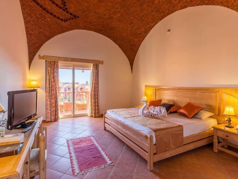 Helles Schlafzimmer mit Holzmöbeln, Doppelbett, Fernseher und Blick auf Balkon mit Glasfenstern.