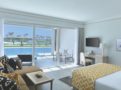 Helles Hotelzimmer mit Doppelbett, Sofa, Schreibtisch und Terrasse mit Blick auf Wasser und Palmen.