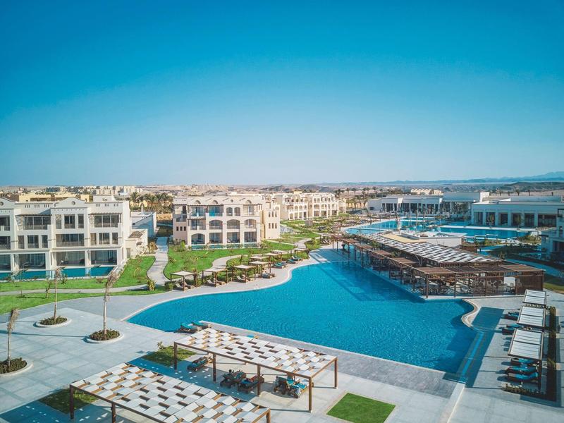 Resort met een groot zwembad, ligbedden en moderne gebouwen onder een helderblauwe lucht.