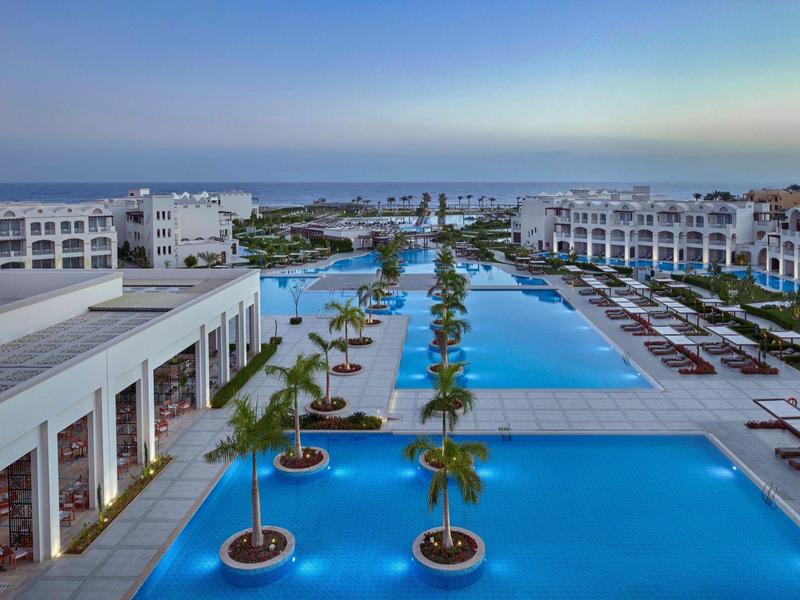 Luxe resort met grote zwembaden, palmbomen, witte gebouwen en uitzicht op de oceaan bij zonsondergang.