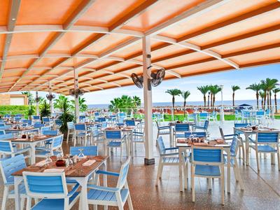 Freiluftrestaurant mit blauen Stühlen und weißen Tischen, Dachschatten und Blick auf Palmen und Meer.
