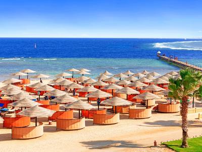Strand mit vielen Sonnenschirmen, Sand, Palmen und blauer See unter klarem Himmel.