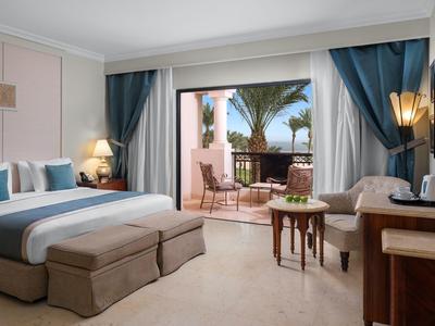 Modern hotel room with double bed, balcony, and view of palm trees and sea.