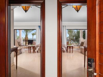Open wooden doors lead to a bright room with furniture and a terrace view with palm trees.