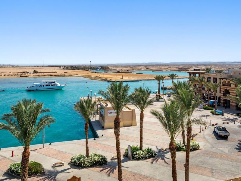 Widok na marinę z palmami, samochodem, łodziami i budynkami pod jasnym niebem.