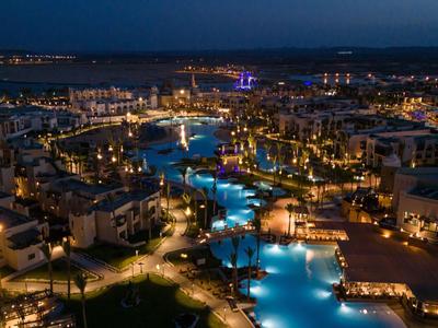 Nachtaufnahme eines beleuchteten Resortpools mit umliegenden Gebäuden und Meer im Hintergrund