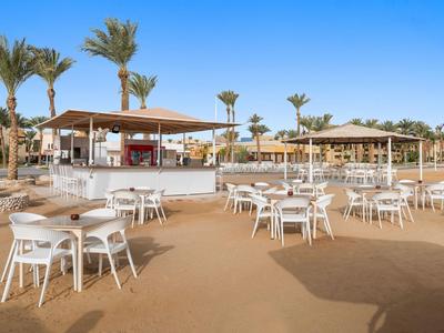 Außenbereich eines Hotels mit weißen Tischen und Stühlen auf Sand unter blauem Himmel.