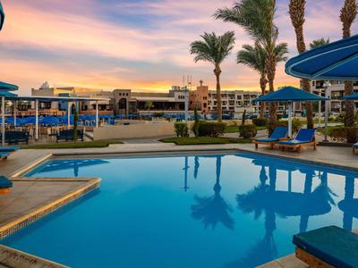 Hotelpool mit Sonnenliegen und blauen Sonnenschirmen bei Sonnenuntergang.