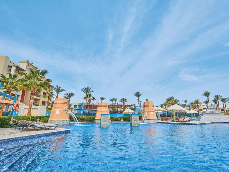 Großes Schwimmbecken mit klar blauem Wasser, umgeben von Palmen und Gebäuden unter blauem Himmel.