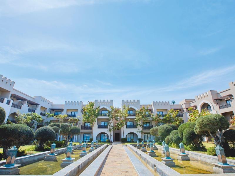 Großes weißes Gebäude mit Burgtürmen, umgeben von symmetrischen Gärten, Brunnen und blauem Himmel.