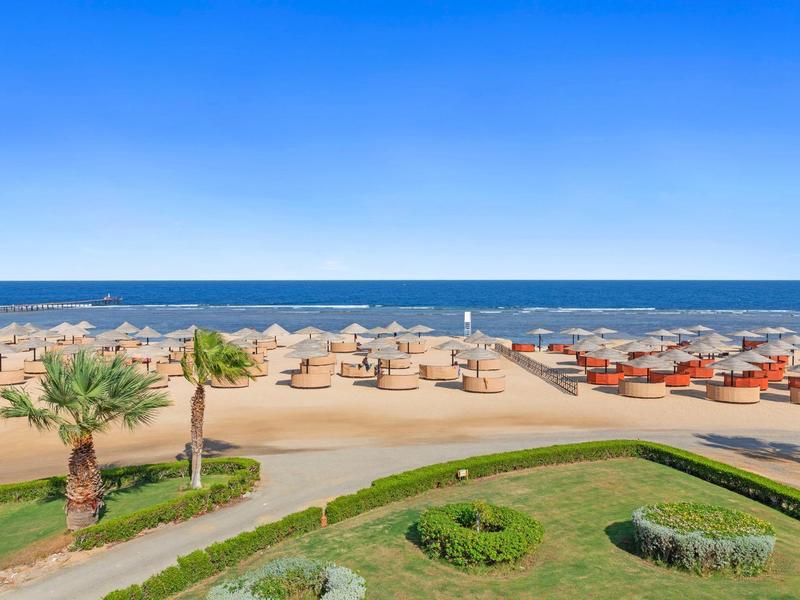 Strand mit Sonnenschirmen, Palmen und blauem Meer unter klarem Himmel.