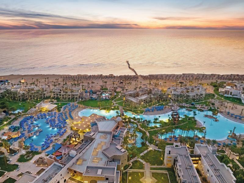 Große Hotelanlage mit mehreren blauen Pools und Palmen an einem Strand bei Sonnenuntergang.