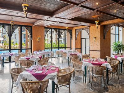 Světla jídelna s kulatými stoly, červenými ubrusy a velkými okny.