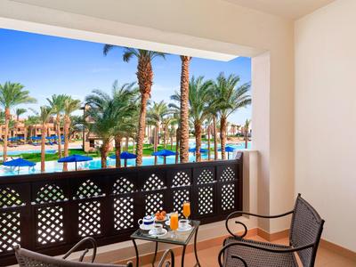 Hotelová terasa se stolem a židlemi, výhled na bazén a palmy pod modrou oblohou.