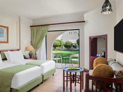 Světelný hotelový pokoj se zelenými dvojlůžky a terasou s výhledem do zahrady.